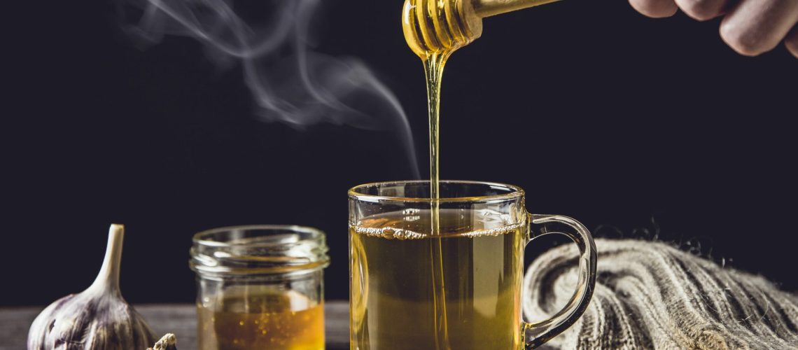 Man hand holding wooden honey dipper, honey spoon on top of glass of tea/ medicine and dripping honey in hot tea. Knitted socks, small jar of honey, garlic on wooden table against black background.