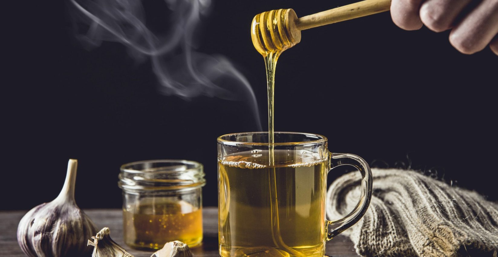 Man hand holding wooden honey dipper, honey spoon on top of glass of tea/ medicine and dripping honey in hot tea. Knitted socks, small jar of honey, garlic on wooden table against black background.