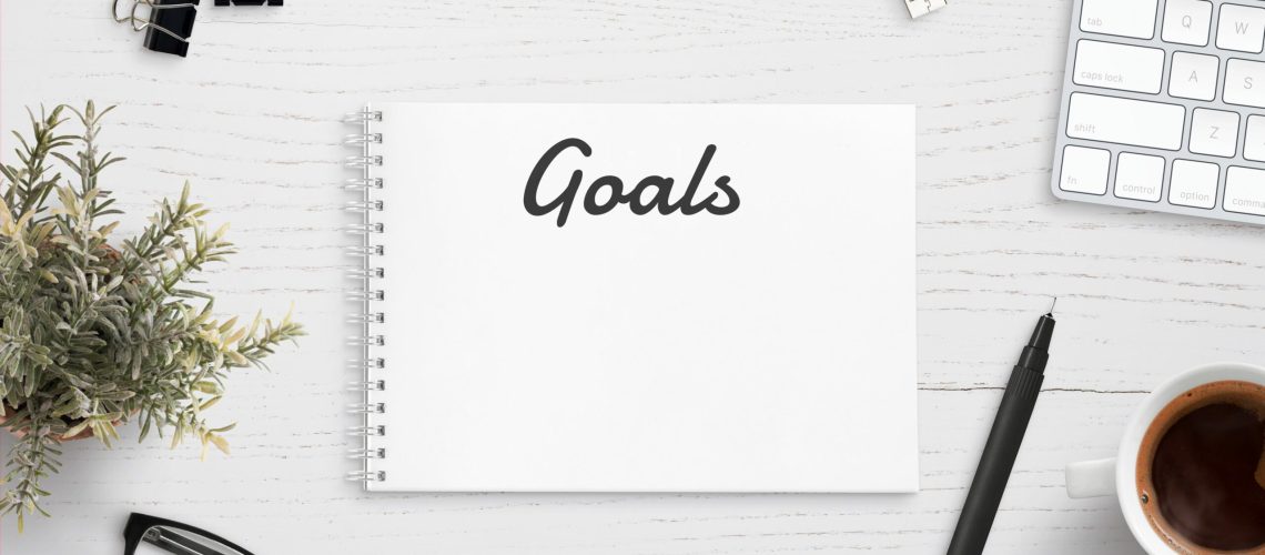 Creating goals list on notepad on office desk surrounded with office supplies. White wooden work desk.