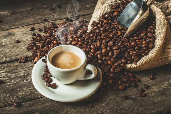 steaming coffee in traditional ceramic cup, jute sack with roasted beans on wooden table.