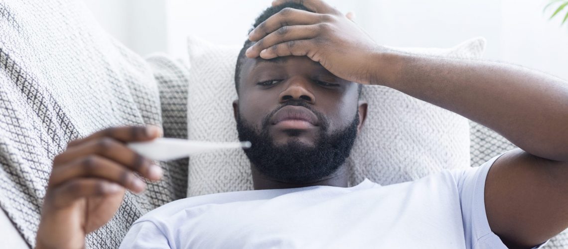 Must be sick. Sad african-american guy with fever measuring his body temperature, lying on couch at home